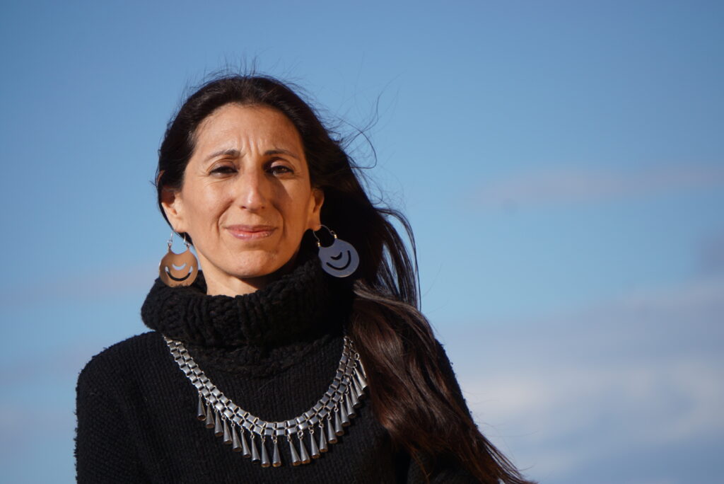 Fotografía de la autora, vestida con un pulóver de lana negra con cuello tipo polerón, con su platería mapuche, con el pelo suelto cayendo sobre su hombro izquierdo, mirando a cámara con una media sonrisa, con el fondo del cielo celeste