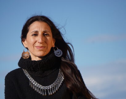 Fotografía de la autora, vestida con un pulóver de lana negra con cuello tipo polerón, con su platería mapuche, con el pelo suelto cayendo sobre su hombro izquierdo, mirando a cámara con una media sonrisa, con el fondo del cielo celeste