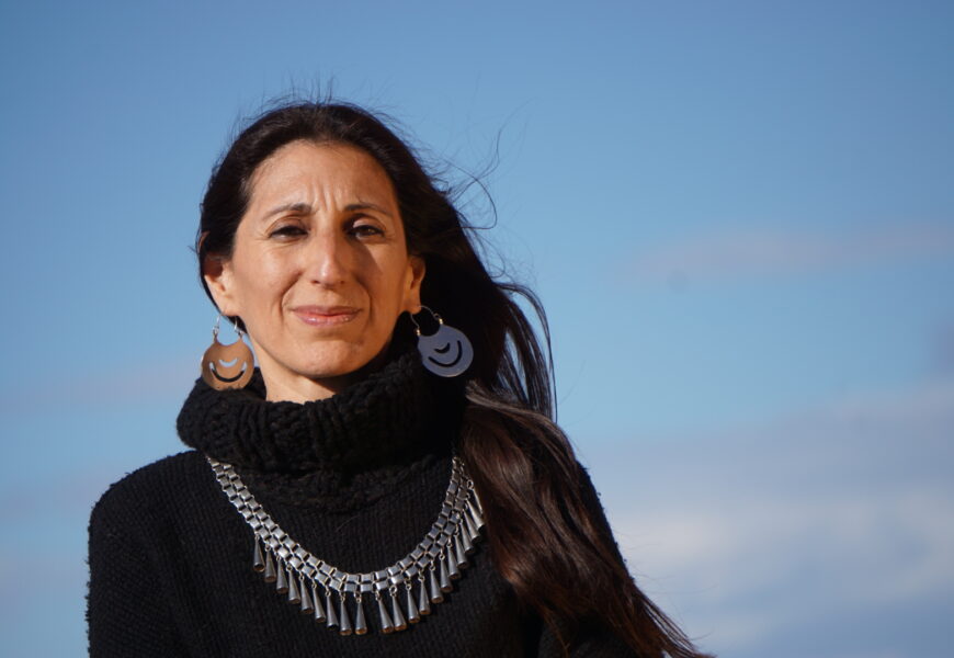 Fotografía de la autora, vestida con un pulóver de lana negra con cuello tipo polerón, con su platería mapuche, con el pelo suelto cayendo sobre su hombro izquierdo, mirando a cámara con una media sonrisa, con el fondo del cielo celeste