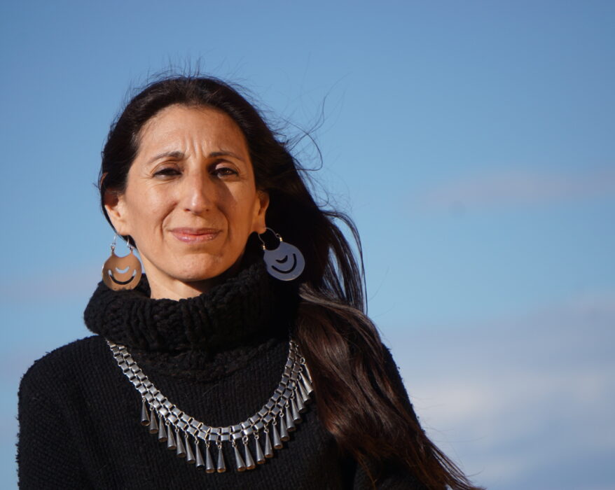 Fotografía de la autora, vestida con un pulóver de lana negra con cuello tipo polerón, con su platería mapuche, con el pelo suelto cayendo sobre su hombro izquierdo, mirando a cámara con una media sonrisa, con el fondo del cielo celeste